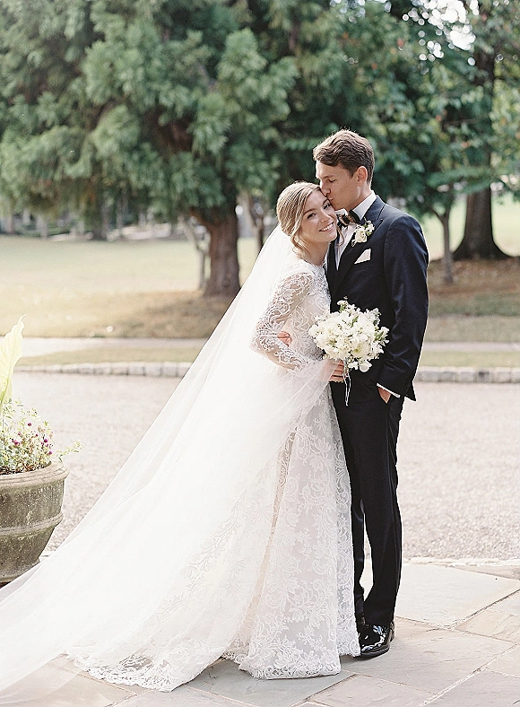 Couple portrait of groom kissing bride’s forehead as she holds a white bouquet, long veil trailing beside trees and stone walkway