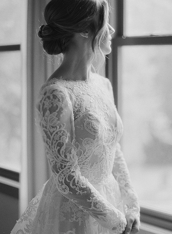 Bridal portrait of a bride looking out window in profile, lit by soft window light, wearing a lace wedding dress with long sleeves and updo