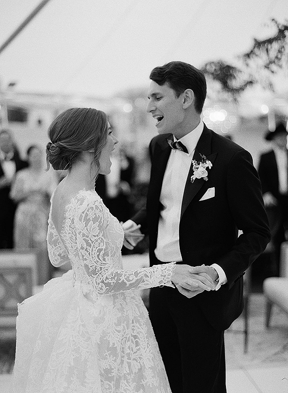 First dance as bride in a lace wedding dress twirls with tuxedo groom under string lights at an outdoor reception with guests behind