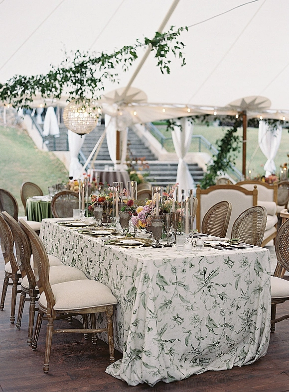 Reception tablescape in a wedding tent reception with patterned linen, wildflower centerpiece, taper candles, and rattan chairs under string lights