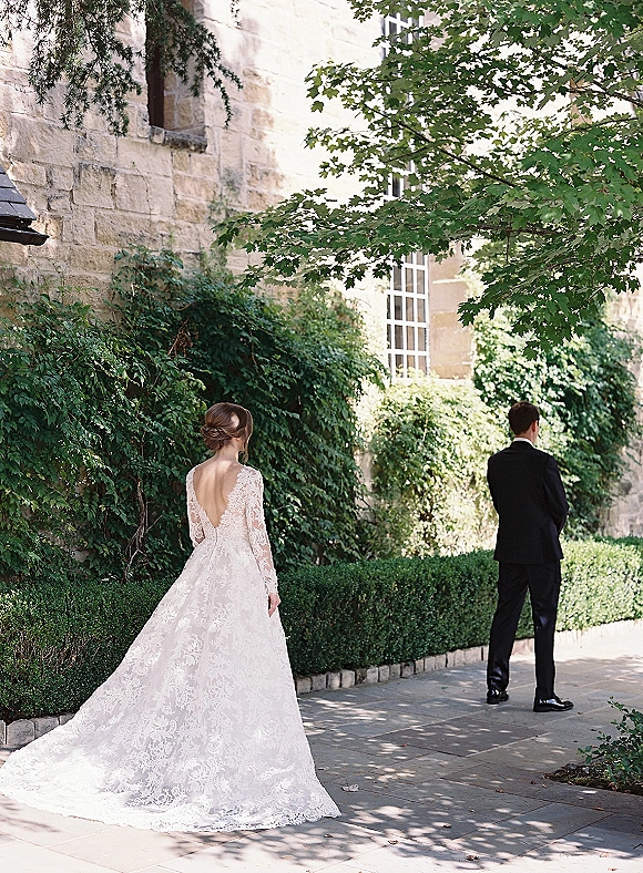 Wedding first look as bride walking to groom in a long sleeve backless lace gown with train on a paved courtyard by ivy stone wall