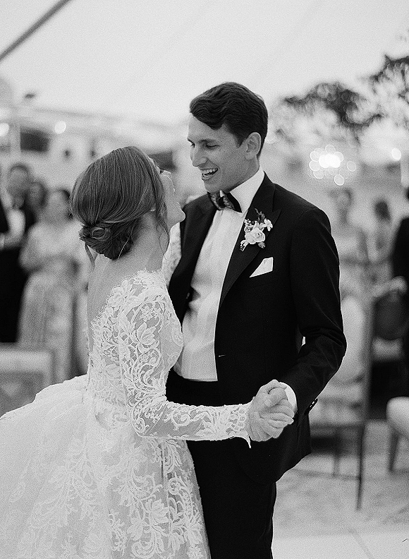 First dance of bride in lace long sleeve dress and groom in tuxedo under string lights, guests watching on an outdoor evening dance floor