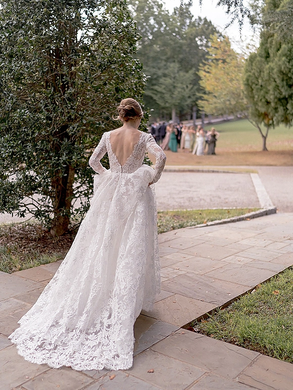 Bridal portrait of a bride from behind holding her lace long-sleeve gown’s cathedral train on a stone walkway in a tree-lined park