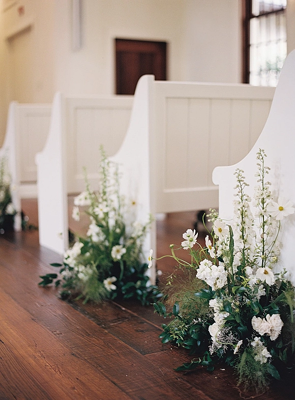Ceremony aisle decor with wedding aisle flowers in white florals and greenery, clustered along a wood floor between white church pews