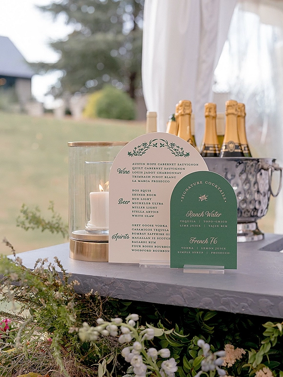 Wedding bar signage and wedding bar menu on a linen table with champagne in an ice bucket, hurricane candle, and greenery under a draped tent on a lawn