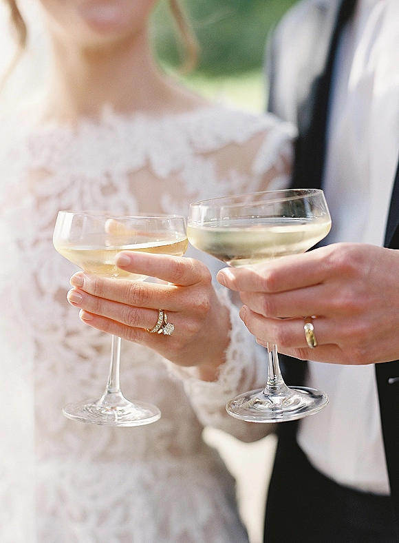 Wedding toast as bride and groom clink champagne coupe glasses, rings visible beside lace sleeve and tuxedo against blurred greenery backdrop