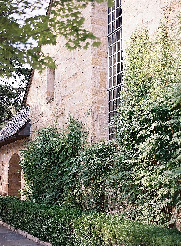 Stone chapel exterior with ivy-covered walls and slate roof, featuring a tall grid window and trimmed hedges along the sidewalk path