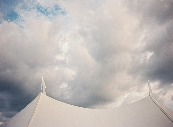 Wedding tent with sailcloth peaks and tall poles, accented by pennant flags beneath a dramatic cloudy sky with patches of blue