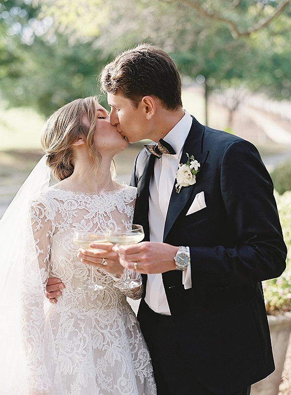 Wedding kiss portrait of bride and groom kissing while holding champagne coupes, lace gown and black tuxedo, sunlit garden bokeh behind