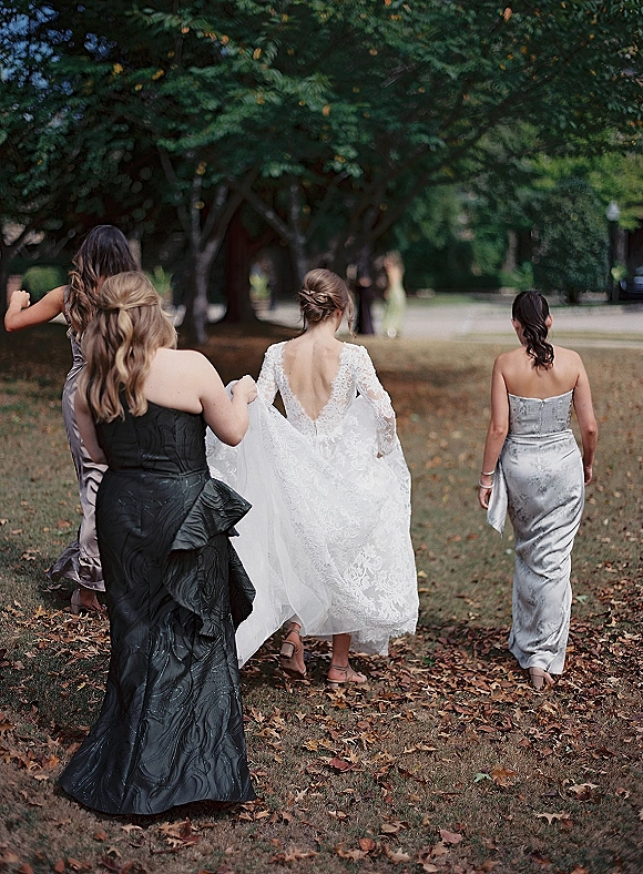 Bride walking from behind in a backless lace wedding dress as bridesmaids carry her train along a park path with fallen leaves