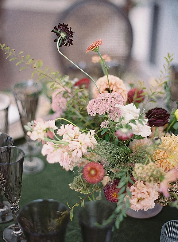 Wedding centerpiece with garden wedding centerpiece blooms and greenery in a bowl, surrounded by smoked goblets and plates on green linen outdoors
