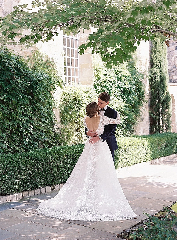 Wedding kiss portrait of bride and groom kissing, her backless lace gown and long train by an ivy-covered stone building courtyard