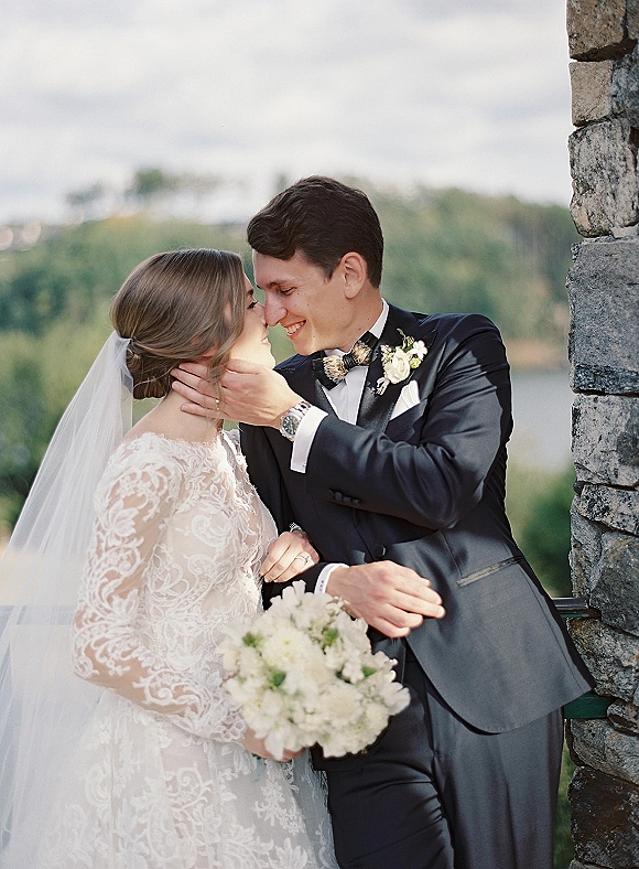 Wedding couple portrait with bride and groom close embrace, veil and white bouquet as he cradles her face by a stone wall and lake