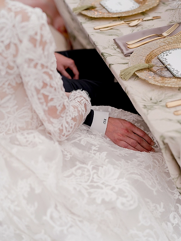 Reception tablescape with gold flatware place setting, woven charger plates, menu card, tassel napkins, and couple’s hands at the table
