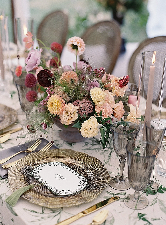 Reception tablescape with wedding table centerpiece of colorful florals in a compote bowl, taper candles, gold flatware, and woven chargers outdoors