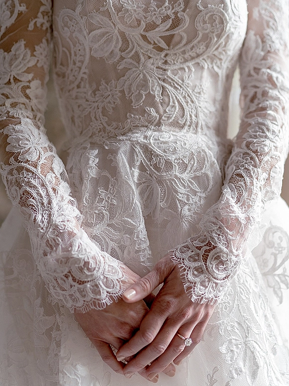 Wedding dress detail showing lace wedding dress sleeves with scalloped cuffs, manicured hands and engagement ring in soft indoor light