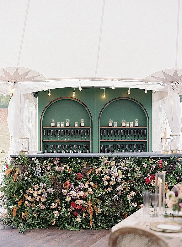Wedding bar setup with a floral bar front and green arched shelving, champagne flutes and candlelight under string lights in a white tent