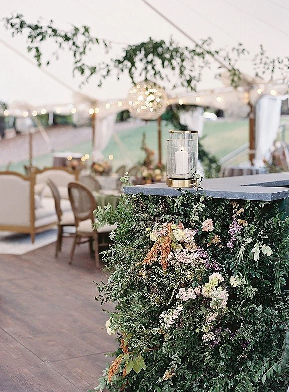 Wedding bar decor with greenery wrapped bar, floral arrangement and lantern candle on the counter under a clear top tent with string lights