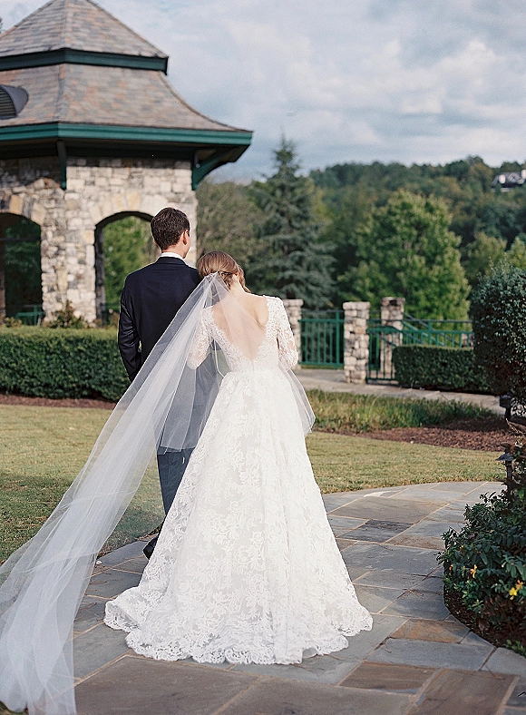 Couple portrait with bride and groom from behind, lace gown and long veil trailing on stone pavilion patio with hills beyond