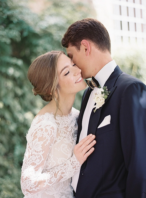 Wedding couple portrait of groom kissing bride’s forehead as she smiles in a lace long sleeve dress, embraced in a greenery garden background