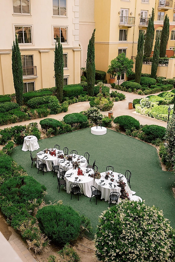 Outdoor reception setup with round tables in white linens and black chairs, greenery centerpieces and candles in a garden courtyard with cypress trees