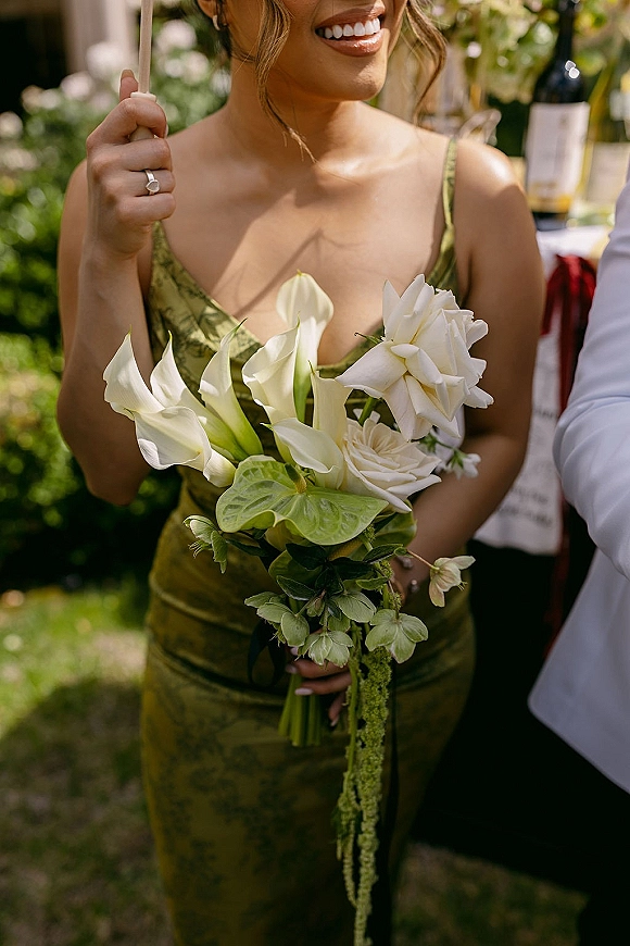 Bridesmaid bouquet of white calla lilies and roses with green anthurium accents held by a woman in an olive spaghetti-strap dress on a lawn