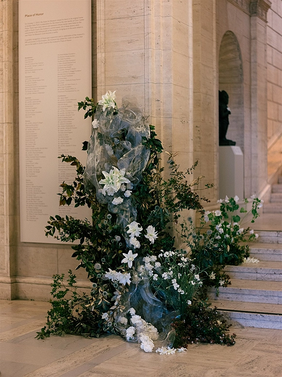 Wedding floral installation of white flowers and greenery with sheer fabric draping at a stone staircase entrance in an arched corridor