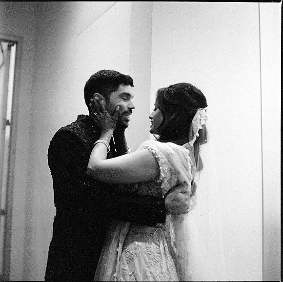 Couple portrait in black and white wedding portrait style, bride holding groom’s face as they embrace indoors by a window frame