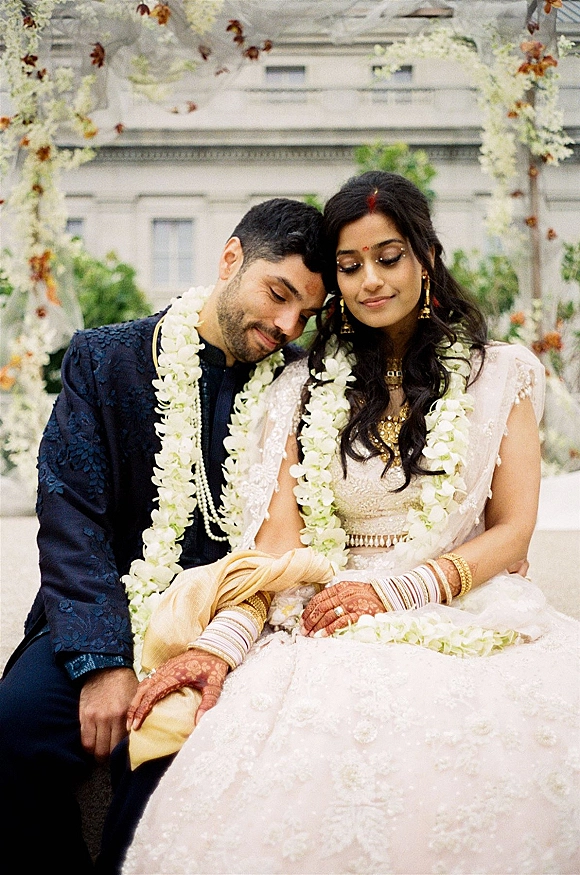 Couple portrait of an Indian wedding couple seated under an outdoor mandap, wearing flower garlands with hanging florals behind them