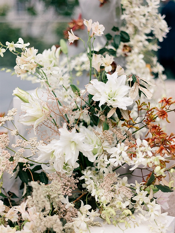 Wedding floral arrangement with white lilies and orchids, greenery accents and orange blooms beside an outdoor walkway lined with plants