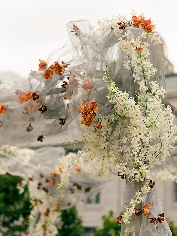 Wedding arch florals with an orchid wedding arch design, featuring white and orange orchids and sheer tulle, set outdoors by greenery