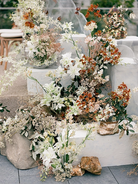 Ceremony altar decor with grounded floral altar of white lilies, rust-orange blooms, baby’s breath and draped fabric on stone blocks in a garden patio