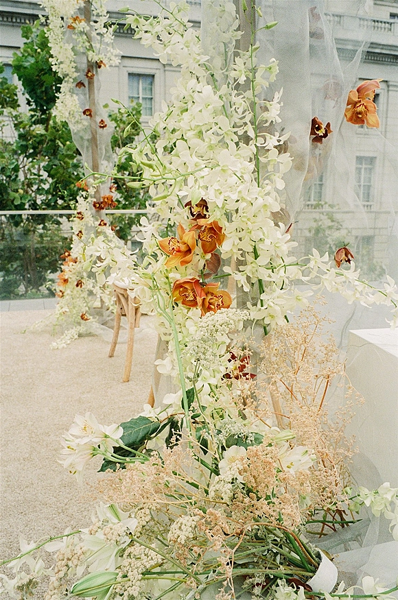 Wedding floral arch with cascading white orchids and sheer draping in a garden courtyard beside a stone building facade and trees