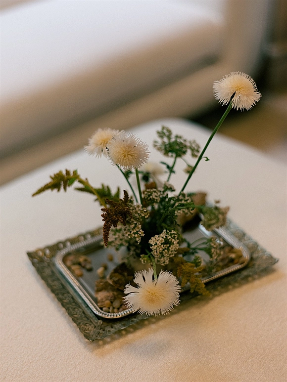 Wedding floral centerpiece with wildflowers and greenery in a small bud vase on a silver tray atop a linen tablecloth, blurred interior behind