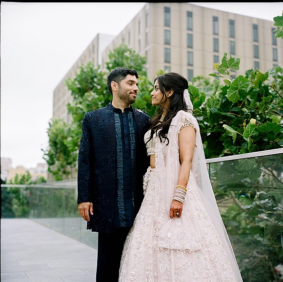 Couple portrait of a south asian wedding couple in lehenga and sherwani, gazing at each other on a rooftop terrace by a glass railing.