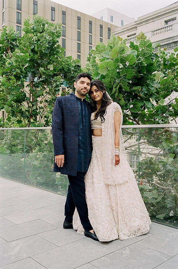 Couple portrait of an Indian wedding couple in blush lehenga and navy sherwani on a rooftop terrace with glass railing and city skyline