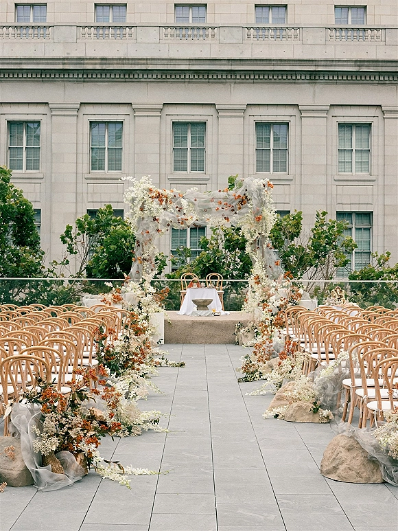 Ceremony setup with an outdoor ceremony aisle leading to a floral ceremony arch with white and orange blooms, draped fabric, and wooden chairs on a terrace