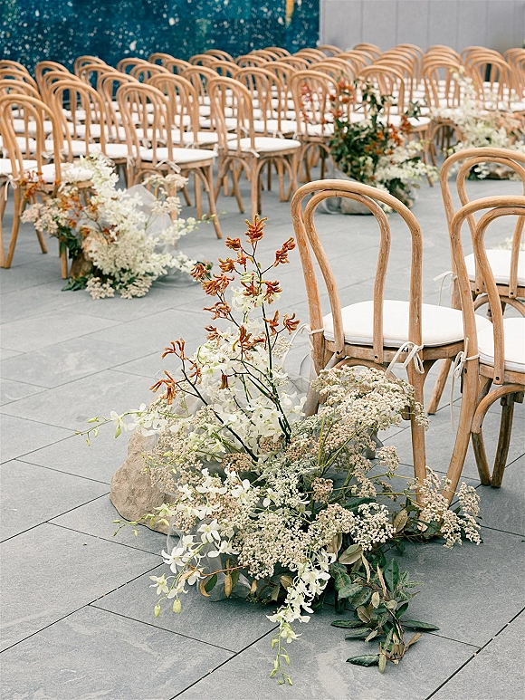 Ceremony aisle decor with wedding aisle flowers lining bentwood chairs, grounded florals and greenery on tile floor beside a dark wall