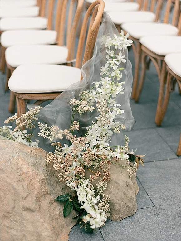 Ceremony aisle flowers with white blooms and greenery arranged low beside wooden chairs, with tulle drape over stone tile aisle floor