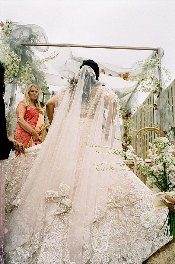 Bridal entrance as bride in embroidered lehenga and veil walks down an outdoor aisle under a white floral arch with chairs and skyline behind