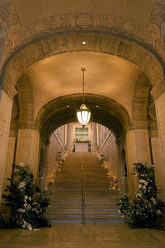 Ceremony aisle design with candlelit staircase wedding ceremony, lined with white florals and greenery beneath an ornate stone archway