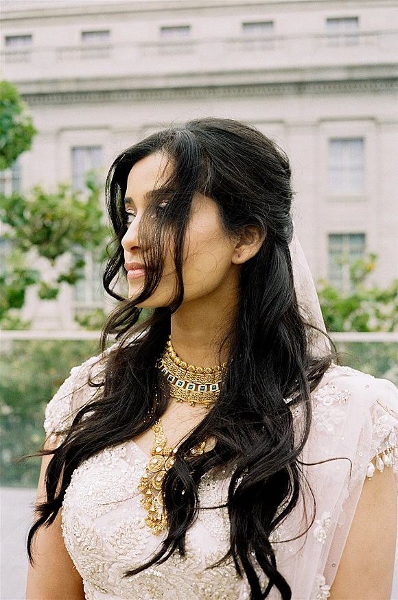 Bridal portrait of a bride in side profile with a bridal veil and gold necklace, looking away beside a historic facade and greenery.