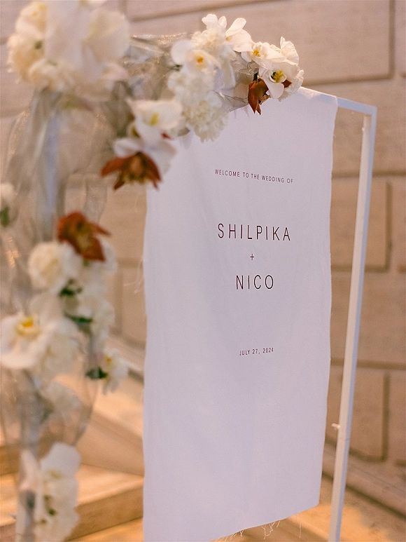 Wedding welcome sign on a metal stand, white acrylic with orchid floral garland and tulle drape, set by stone steps and wall