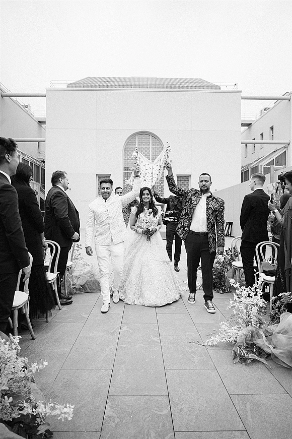 Wedding recessional as bride walking down aisle with bouquet and veil beside groom in patterned jacket and sneakers on outdoor terrace with cheering guests