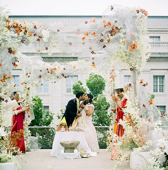 Ceremony kiss at a wedding ceremony moment beneath a draped floral arch with white and orange flowers on an outdoor terrace backdrop