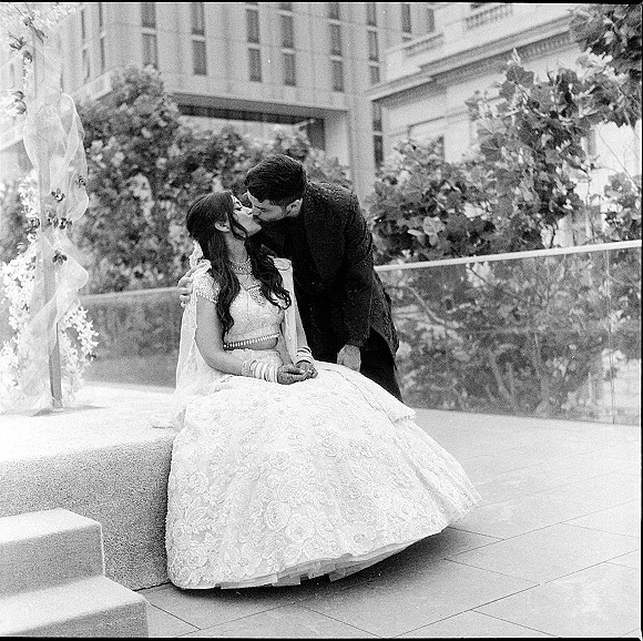 Wedding kiss portrait of a bride and groom kissing while seated on stone steps, her embroidered lehenga and dupatta beside his sherwani on a terrace