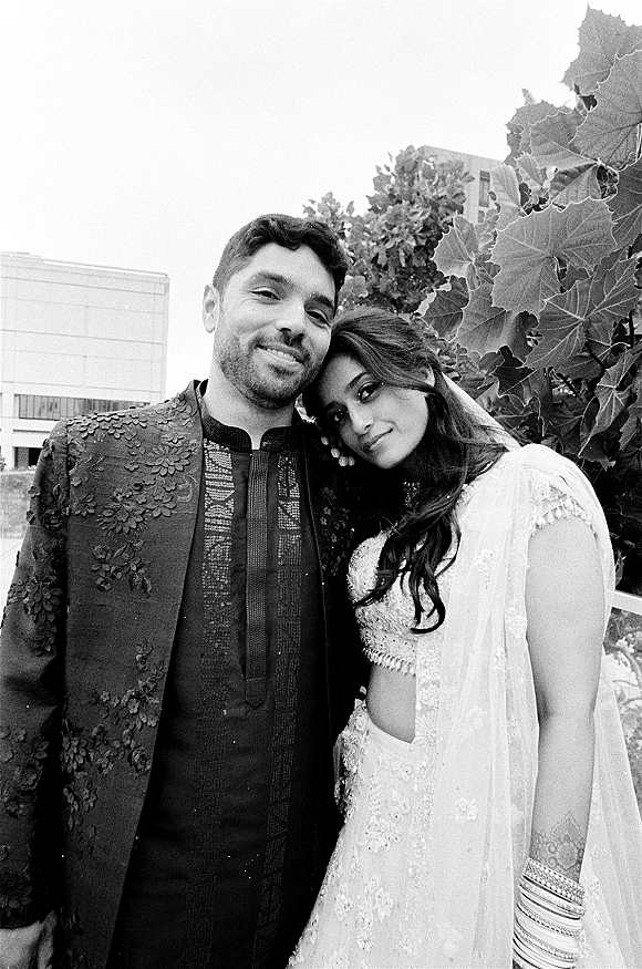 Couple portrait in a black and white wedding portrait, bride leaning on groom in sherwani and lehenga on a leafy terrace backdrop
