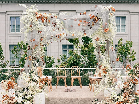 Wedding ceremony arch with sheer draping and white flowers, accented by orange blooms, flanked by wooden chairs before a building facade