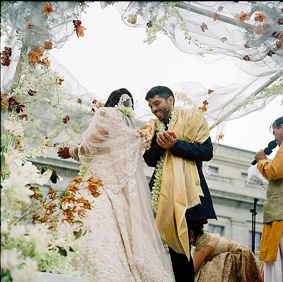 Ceremony moment at a Hindu wedding ceremony as bride places floral garland on groom under a draped mandap canopy with hanging flowers outdoors