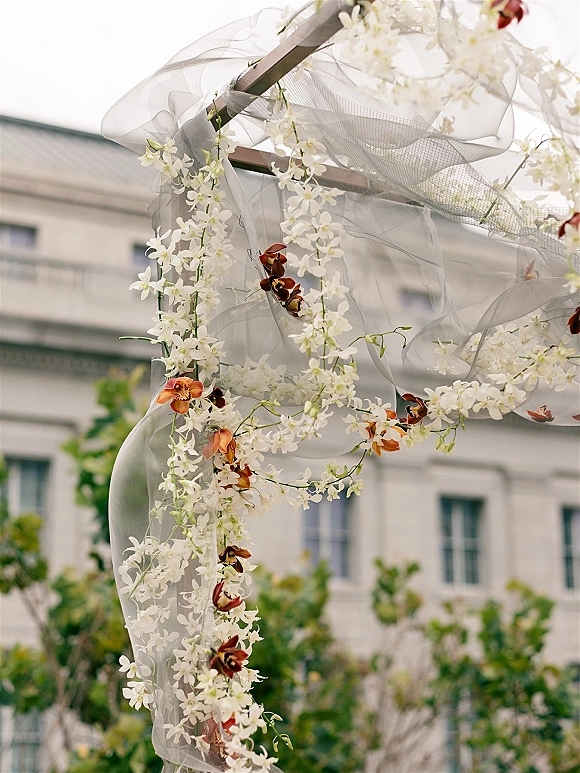 Wedding ceremony arch with white orchid garlands and sheer drapery on a metal frame, set outdoors near a venue building and greenery